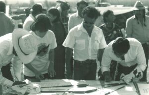 People sign large documents on a table outdoors at a group event, with onlookers in the background (black-and-white photo).
