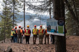 Group of hikers wearing orange safety vests and helmets reading a bat habitat information sign in a wooded clearing in the mountains form a line in front of the sign.