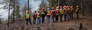 Group of workers in neon safety vests and hard hats stand in a forest clearing, listening to a guide for a safety briefing on a rocky hillside.