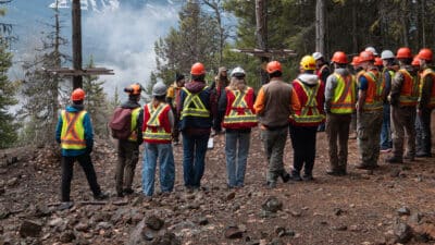 Group of workers in neon safety vests and hard hats stand in a forest clearing, listening to a guide for a safety briefing on a rocky hillside.