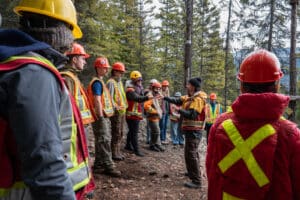 Group of people in orange and yellow safety vests and helmets standing in a forest, listening to a speaker during a training drill.