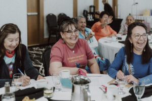 Group of smiling adults seated around a table during a conference or lunch event, cups and papers in front of them.