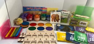 Colorful preschool learning shelf with color-sort containers, paint jars, wooden number and color mats, alphabet cards, animal flashcards, and numbered dice for early math