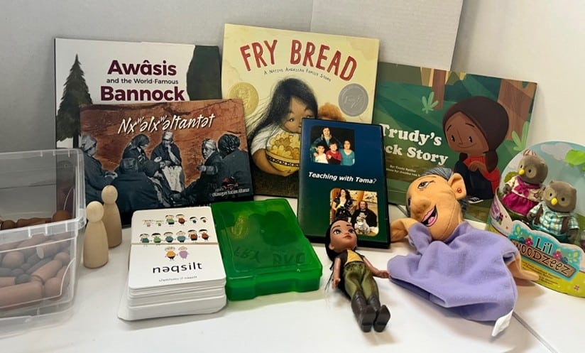 Assorted children's books, teaching cards, and toys on a shelf: titles include Fry Bread and Awáis Bannock, plus a green storage box, small figures, and a purple-clothed doll.