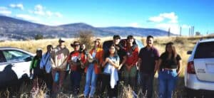 Group of diverse young people posing for a photo between two parked SUVs in a dry field with mountains in the distance.