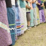 Row of people standing in a line outdoors, each wearing vibrant patterned long skirts and crossing their arms.