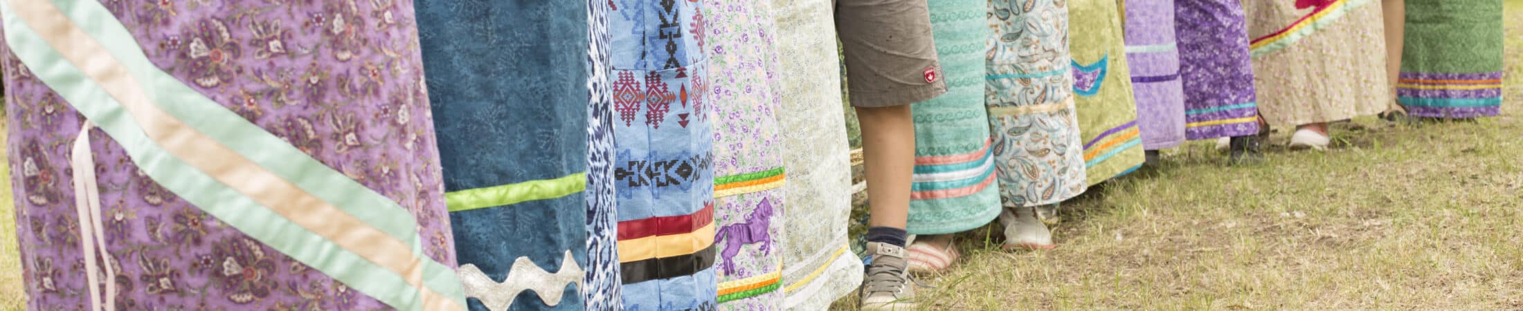 Row of people standing in a line outdoors, each wearing vibrant patterned long skirts and crossing their arms.
