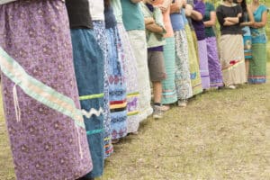 Row of people standing in a line outdoors, each wearing vibrant patterned long skirts and crossing their arms.
