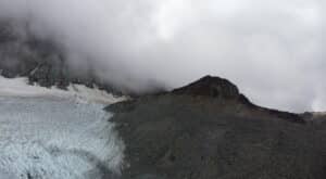 Snow-covered glacier on the left cliffs with a rocky slope on the right, shrouded in low clouds and mist.
