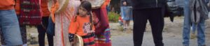 Young girl in an orange shirt holding a small frame drum among a group of people in colorful clothing outdoors.