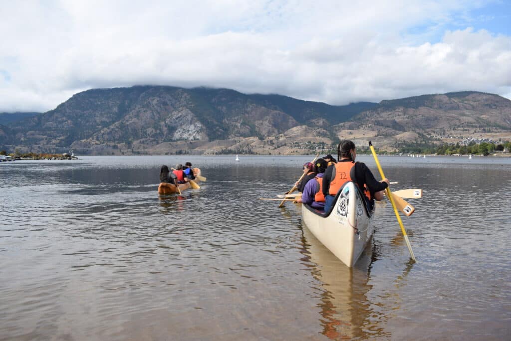 People in orange life jackets paddling a canoe on a calm lake with a mountain range in the background and cloudy sky.