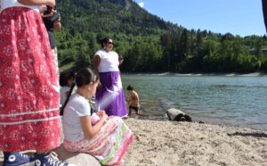 Group of children and adults in colorful clothing standing on a rocky lakeshore, with a forested mountain and blue sky behind them.