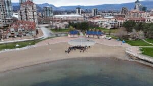 Drone shot of a waterfront park with a group of people on a sandy beach, blue-roofed kiosks, and city buildings in the background.