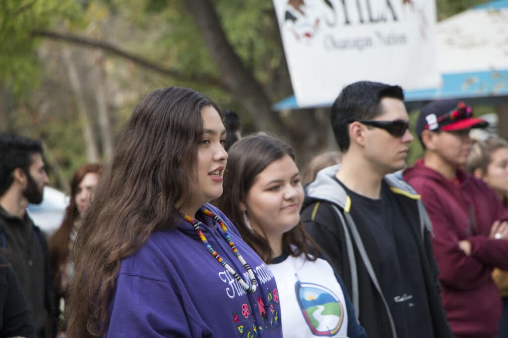 Woman foreground wearing a purple hoodie and colorful beaded necklace, standing in a crowd outdoors with a banner in the background.