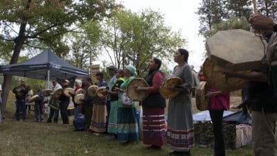 Group of people standing in a line outdoors, playing round drums as part of a cultural performance near a tent.
