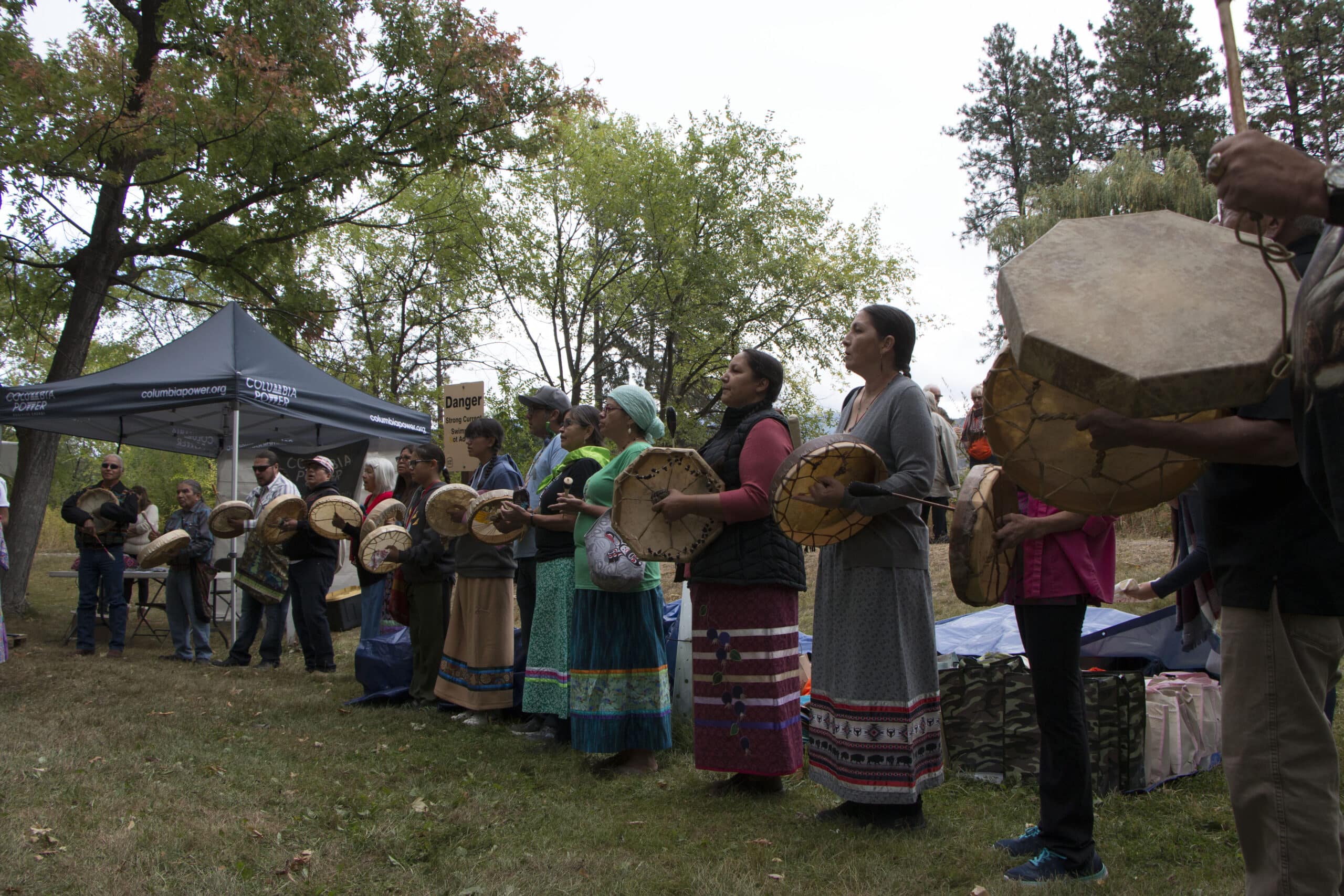 Group of people standing in a line outdoors, playing round drums as part of a cultural performance near a tent.