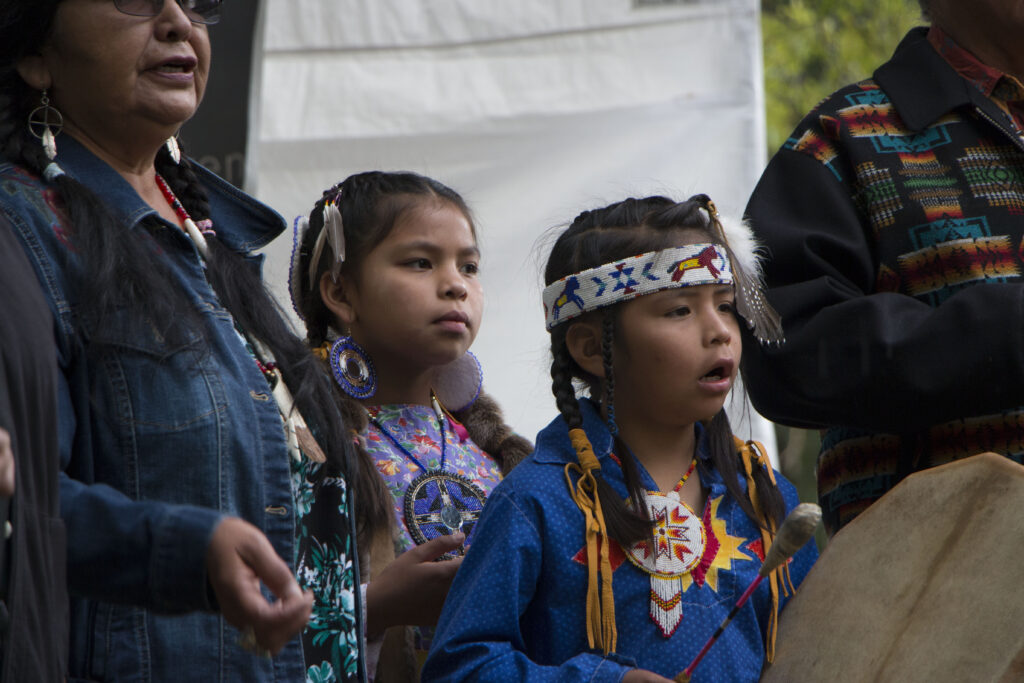 Two Indigenous girls in colorful regalia and an adult woman in a denim jacket, standing outdoors near a horse at a cultural event.
