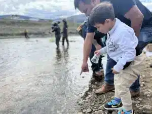 Children and adults crouch at a rocky shoreline, bending toward shallow water and pointing at the surface while a clear plastic cup is in a child's hand.