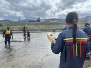 Person with braided hair in a denim jacket with colorful ribbons stands near a river, with people in life jackets and a rescue drill in the background.