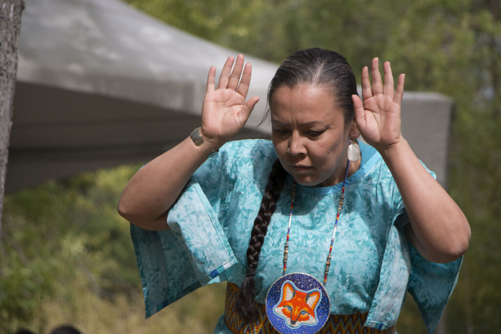 Woman in turquoise clothing with braided hair raises both hands in front of her, outdoors in a grassy area.