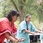 Two women in bright traditional outfits perform a dance outdoors while a crowd watches under trees in a park-like setting.