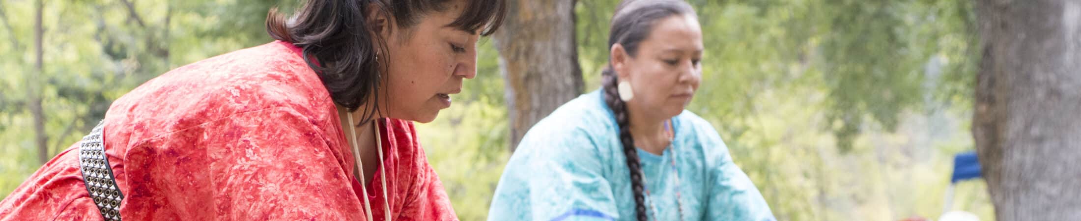 Two women in bright traditional outfits perform a dance outdoors while a crowd watches under trees in a park-like setting.