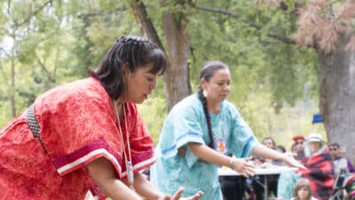 Two women in bright traditional outfits perform a dance outdoors while a crowd watches under trees in a park-like setting.