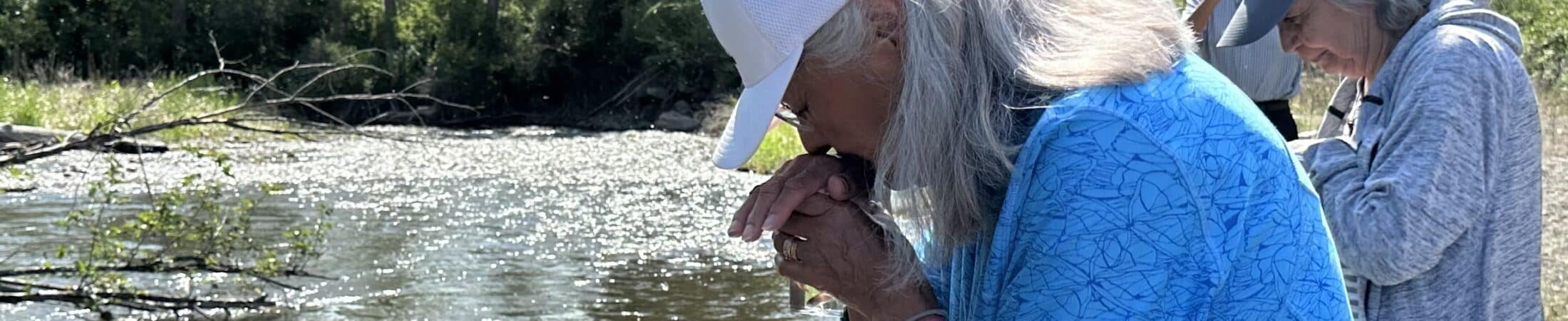 Two older women stand at a sunlit riverbank, looking down at the water; one wears a white cap and bright blue patterned shirt, the other a gray cap and jacket, with trees and power lines in the background.
