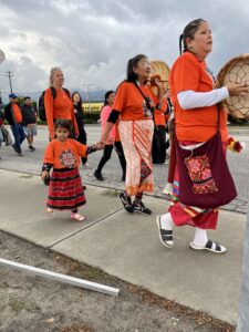 People participate in a street parade wearing bright orange shirts; a child in an orange top and red skirt holds an adult's hand in the foreground.