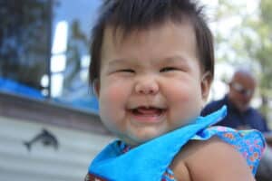 Close-up of a smiling baby with a blue towel around the shoulders, outdoors on a sunny day.