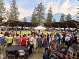 Large crowd gathered outdoors at a festival with wooden shade structures and a mountain backdrop under a cloudy sky.