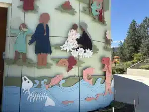 Outdoor wall mural with layered paper-cut silhouettes of people and dinosaurs in pastel colors, atop a blue water-themed base.