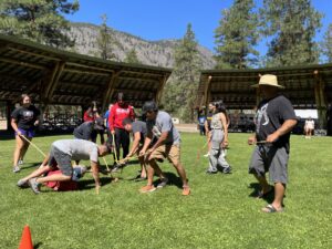 Group of people outdoors on a grassy field playing a game with sticks, orange cones, and a sheltered pavilion nearby. Mountains and trees rise in the background.