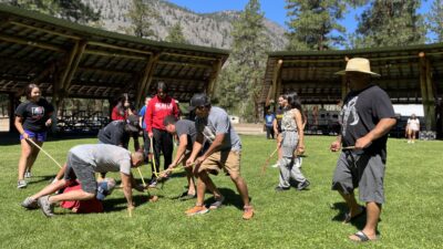 Group of people outdoors on a grassy field playing a game with sticks, orange cones, and a sheltered pavilion nearby. Mountains and trees rise in the background.