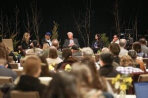 Panel discussion on stage with five panelists seated, audience in foreground, bare trees as backdrop.