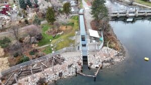 Aerial view of a riverside park showing a long metal ramp and platform over a rocky shoreline, with trees, a walking path, and a colorful play structure in the distance.