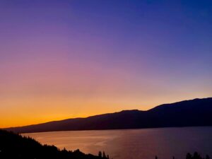 Sunset over a calm lake with orange-to-purple sky and dark mountain silhouettes along the shore.