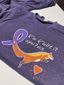 Close-up of a purple t-shirt showing a large orange fox and purple ribbon graphic with white handwritten-style text beside it.