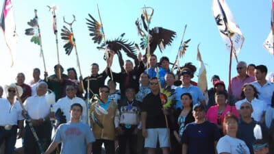 Group of people posing outdoors, each holding tall decorative staffs with colorful feathers and flags during a cultural gathering.